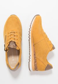 Zapatillas de ante amarillas con textura perforada, detalles de cuero marrón, suelas blancas y un patrón de zigzag en la parte inferior. Incluye cordones y cremallera.