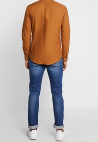 Langarmshirt in gebrandoranje, gemaakt van katoen, met een rugpand met een middenplooi, gecombineerd met blauwe jeans en witte sneakers.