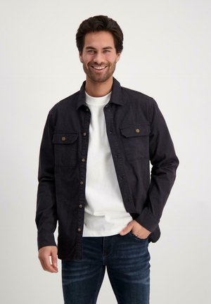 Zwarte overhem shirt met borstzakken, gedragen over een wit T-shirt, gecombineerd met blauwe spijkerbroek. De stof lijkt textuur te hebben met een casual pasvorm.