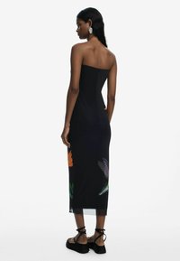 Black strapless midi dress with floral print at the hem. Smooth fabric, fitted silhouette. Model wears black sandals with ankle ties.