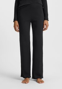 Άτομο που φοράει μαύρο ριμπ παντελόνι φαρδιάς γραμμής και ταιριαστό μακρυμάνικο τοπ, στέκεται ξυπόλητο σε απλό φωτεινό φόντο.