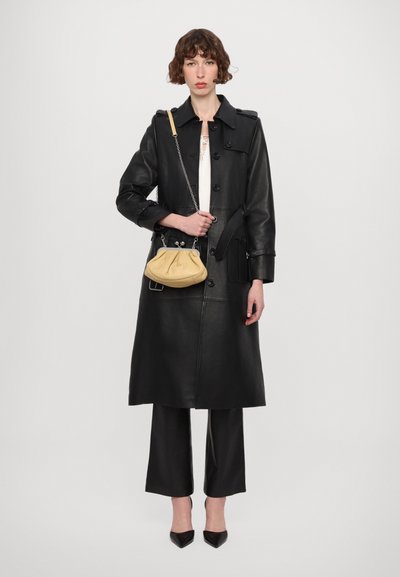 Sort læder trenchcoat med taljebælte, lange ærmer og knaplukninger. Modellen holder en sennepsgul taske med kædelukning.
