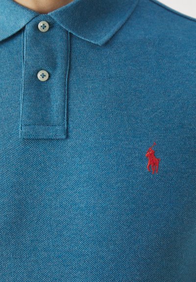 Camisa polo de tejido azul texturizado con un color clásico, tres botones y un pequeño logo bordado en rojo en el lado izquierdo del pecho.