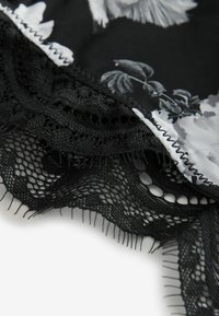 Schwarzer Stoff mit floralem Muster und gewelltem, filigranem schwarzen Spitzenrand mit feinen fransigen Kanten auf einem hellen Hintergrund.