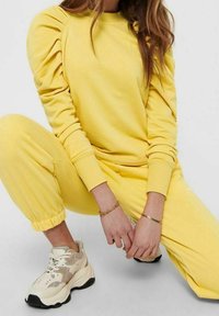 Sudadera amarilla con mangas abullonadas y joggers a juego, hechos de tela suave. Combinado con zapatillas gruesas beige y negras.