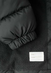 Manche d'une veste en duvet noire avec poignet élastique reposant sur un tissu noir texturé, avec une étiquette blanche comportant les champs "Nom" et "Téléphone".