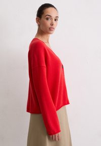 Czerwony kardigan z mieszanki wełny, z dekoltem w serek, długimi rękawami i luźnym krojem; posiada pojedynczy czarny guzik z przodu.