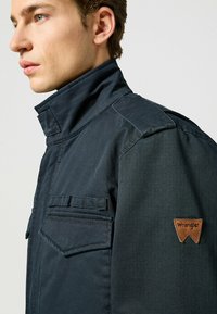 Dunkelblaues Jackett mit hohem Kragen, das eine Brusttasche, Nähte und ein braunes Lederlogo-Patch auf der Schulter aufweist.