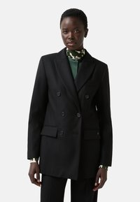 Black double-breasted blazer with notched lapels, decorative buttons, front pockets, and a tailored fit, worn over a green patterned shirt.