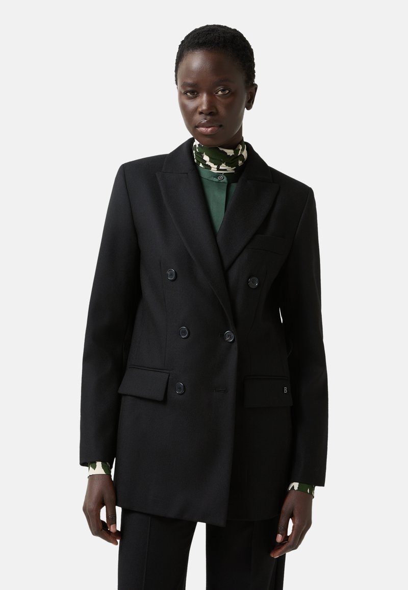 Black double-breasted blazer with notched lapels, decorative buttons, front pockets, and a tailored fit, worn over a green patterned shirt.