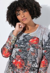 Frau mit lockigem Haar, die das Kinn auf die Hand stützt, trägt ein langärmliges Shirt mit rotem Herz und floralen Mustern vor einem hellen Hintergrund.