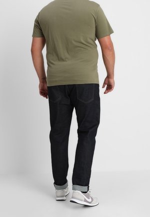 Mężczyzna ubrany w oliwkowozielony t-shirt, ciemne podwinięte dżinsy i białe trampki, stojący tyłem na jednolitym tle.