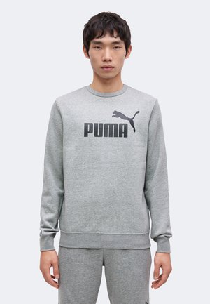 Jeune homme portant un sweat-shirt Puma gris clair et un pantalon de survêtement assorti, debout devant un fond uni.