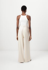 Weißes Tanktop und cremefarbene, weit geschnittene Hose mit hoher Taille, elastischem Bund und zwei Gesäßtaschen, vor einem schlichten Hintergrund.