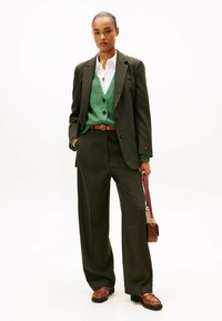 Completo pinstripato verde scuro con pantaloni relax-fit. Sovrapposto a un maglione verde con texture e una camicia bianca. Cintura marrone e borsa coordinata.