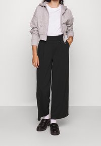 Grå kort hoodie, vit t-shirt, svarta vida byxor med veck och knappar, svarta träskor och ljusa strumpor. Enfärgad bakgrund.