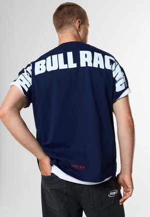 Uomo di spalle che indossa una t-shirt blu navy con grande testo bianco "Red Bull Racing" sulla schiena e pantaloni neri con il logo RBR sulla tasca.