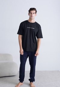 Camiseta de algodón negra con el logotipo blanco "TOMMY HILFIGER", combinada con pantalones de chándal azul marino. El modelo está descalzo sobre una alfombra gris claro.