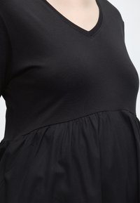 Μαύρο εγκυμοσύνης φόρεμα με V λαιμόκοψη και ραμμένη μέση, που δείχνει τον άνω κορμό και την φουσκωμένη κοιλιά εγκύου μπροστά σε απλό φόντο.