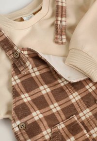 Sweter w kolorze kremowym w połączeniu z brązowymi szelki z korduroyu, zdobionymi wzorem w kratę, guzikami i kieszenią z przodu.