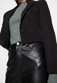 Krótko skrojona czarna marynarka z pojedynczym czarnym guzikiem, zestawiona z teksturowanym zielonym topem oraz wysokim stanem czarnych skórzanych spodni.