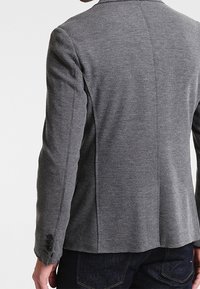 Harmaa bleiseri, jossa on teksturoitu kangas, virtaviivainen muotoilu, kaksinappinen suljenta ja lovilaput, muotoiltu malli ja sivuotsat.