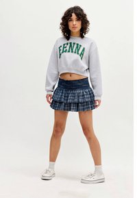 Žena stojící v šedém sportovním svetru s textem "FENNA", v modré kostkované mini sukni s volány, v bílých ponožkách a bílých teniskách na bílém pozadí.