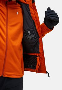 Lyst orange vandtæt jakke med et sort inderfor, der har en lynlås, sidelommer og et indvendigt mærke, der detaljerer teknologien.