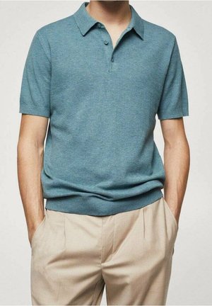 Man draagt een polo met korte mouwen in turquoise met kraag en beige broek, staand met handen in zakken tegen een effen achtergrond.