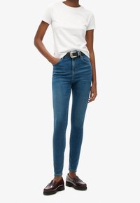 Jeans de mezclilla de corte ajustado en azul medio, con una cintura alta, cinturilla y una hebilla de tono plateado, combinados con una camiseta blanca sencilla.
