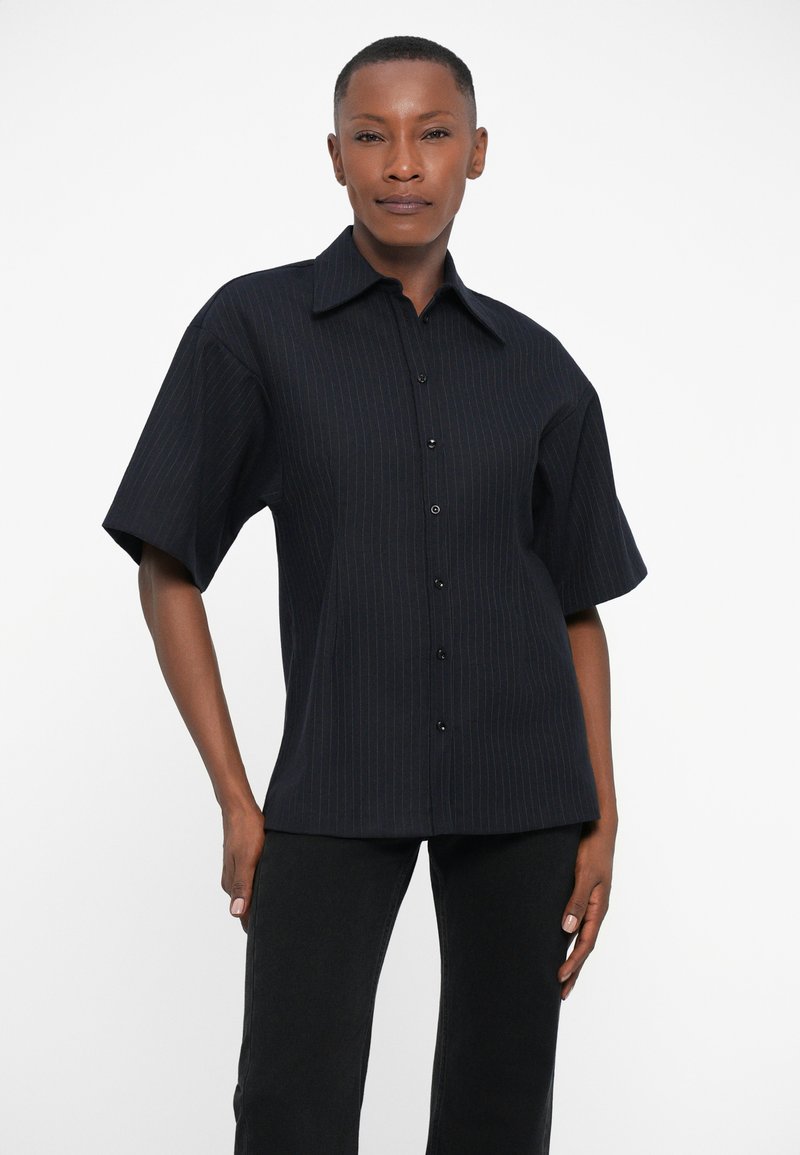 Chemise noire à rayures épaisses à manches courtes avec un col pointu et des boutons sur le devant, fabriquée dans un tissu texturé, associée à un pantalon noir.