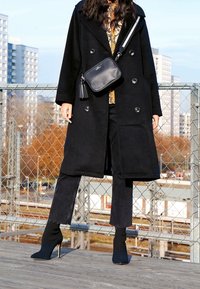 Cappotto nero doppiopetto con bottoni lucidi, abbinato a stivaletti neri con tacco alto e a una piccola borsa nera a tracolla con un pon-pon.