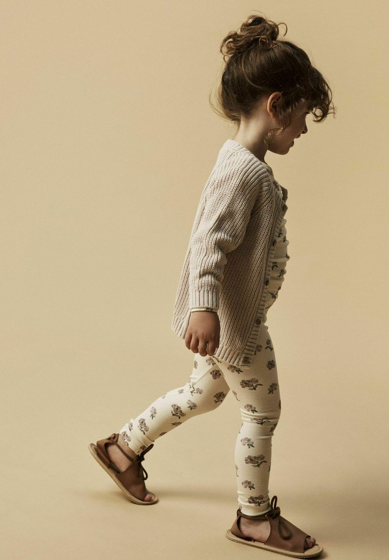 Bambino in un cardigan di maglia beige, leggings floreali color panna e sandali marroni. La texture è rigata e morbida; il design è semplice e decorato.
