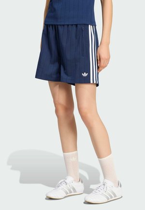 Osoba nosząca granatowe spodenki Adidas z białymi paskami i logo, białe siateczkowe skarpetki oraz białe buty Adidas z szarymi paskami.