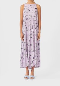 Maxi-jurk van licht lavendelstof met bloemenpatroon, mouwloos, met gelaagde volants aan de onderkant. Gecombineerd met lichtblauwe gevlochten sandalen.