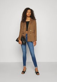 Blazer castanho às riscas, cruzado, combinado com um top preto curto, jeans justos azuis, sandálias pretas de salto e uma mala preta.