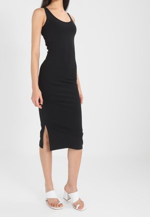 Femme portant une robe midi noire sans manches avec une fente latérale et des sandales blanches à talons blocs ouverts, debout sur un fond blanc uni.