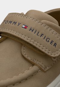 Μπεζ δερμάτινο παπούτσι χωρίς κορδόνια με λουράκι με βέλκρο. Το λογότυπο της μάρκας "TOMMY HILFIGER" εκτυπωμένο σε ναυτικό μπλε. Διαθέτει ραφές και μαλακή υφή.