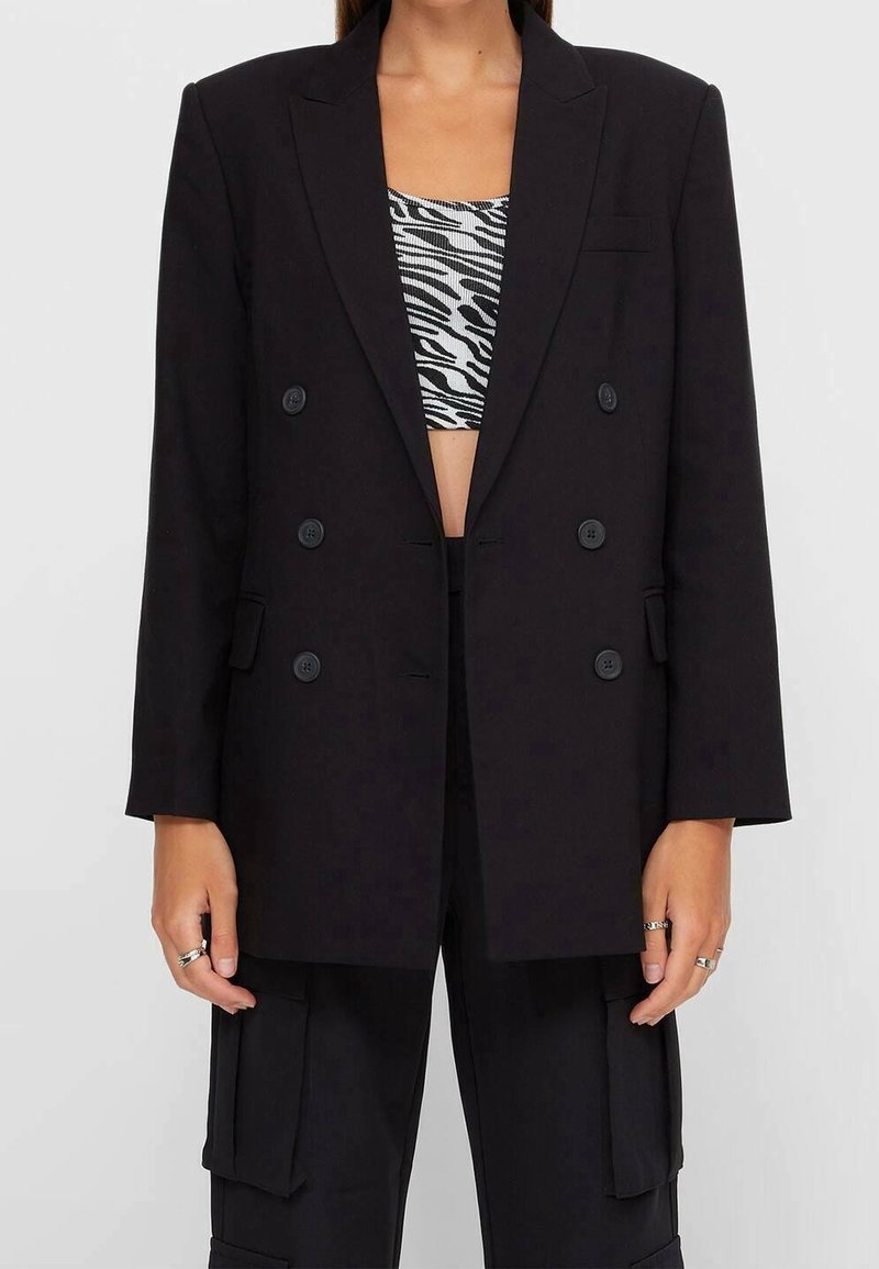 Personne portant un blazer noir oversize à double boutonnage sur un crop top imprimé zèbre noir et blanc et un pantalon cargo noir.