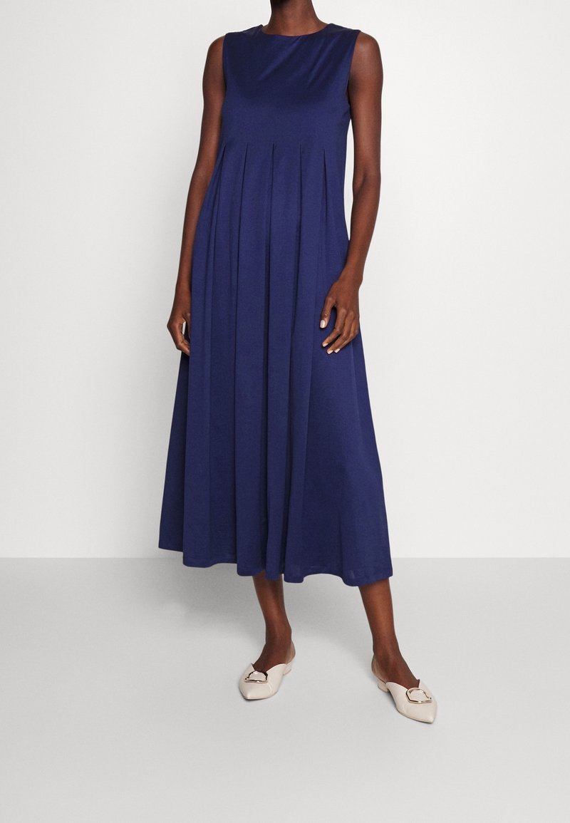 Femme portant une robe midi bleu marine sans manches à plis, associée à des ballerines blanches pointues avec une boucle dorée.