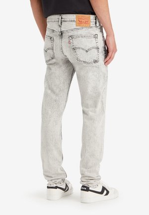 Person, die hellgraue Levi's-Jeans und weiße Sneakers mit schwarzen Akzenten trägt, steht mit dem Rücken zu uns auf einem weißen Hintergrund.