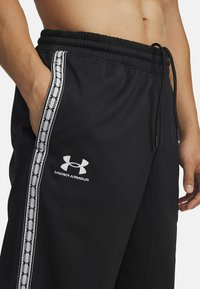 Black athletic pants feature an elastic waistband, side pockets, and a white logo. Side stripes with a repeating pattern add detail.