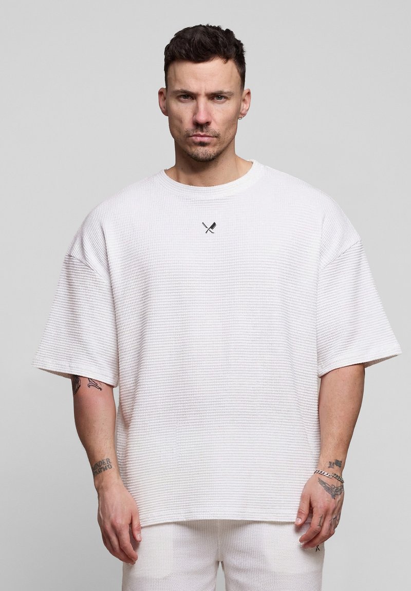 Homme aux cheveux courts et foncés portant un t-shirt blanc texturé surdimensionné et un pantalon, debout devant un fond gris clair uni.