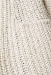 Ljusbeige stickad tröja med ribbat mönster, som har en synlig ficka och mjukt, fluffigt material. Närbild som betonar hantverket.