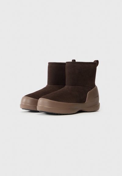 Braune Wildleder-Ankelstiefel mit dicken beigen Gummisohlen, runden Zehen und Zuglaschen auf weißem Hintergrund.
