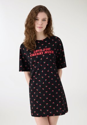 Donna indossa un vestito nero con piccoli motivi a ciliegia e il testo rosso "TI AMO COSÌ TANTO CILIEGIA" sul petto, in piedi contro uno sfondo semplice.