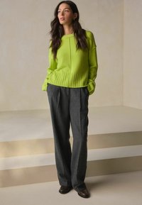 Lime zaļš adīts džemperis ar ribotu apakšmalu, kombinēts ar gaiši pelēkiem, brīvi pieguļošiem, teksturētiem biksēm un tumšām kurpēm. Vienkāršs, mūsdienīgs dizains.