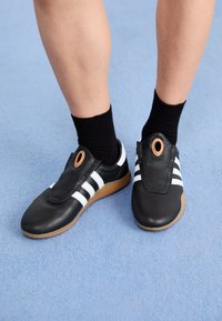 Black slip-on shoes with white stripes and a textured leather upper, featuring a circular accent near the opening and a gum rubber sole.