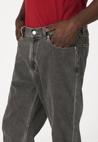 Manos metidas en los bolsillos delanteros de unos vaqueros negros desgastados, combinados con una camisa roja, visto desde la cintura hasta la mitad del muslo.