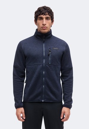 Homme portant une veste zippée bleu foncé texturée avec un col haut et une poche poitrine zippée, debout les bras détendus le long du corps.